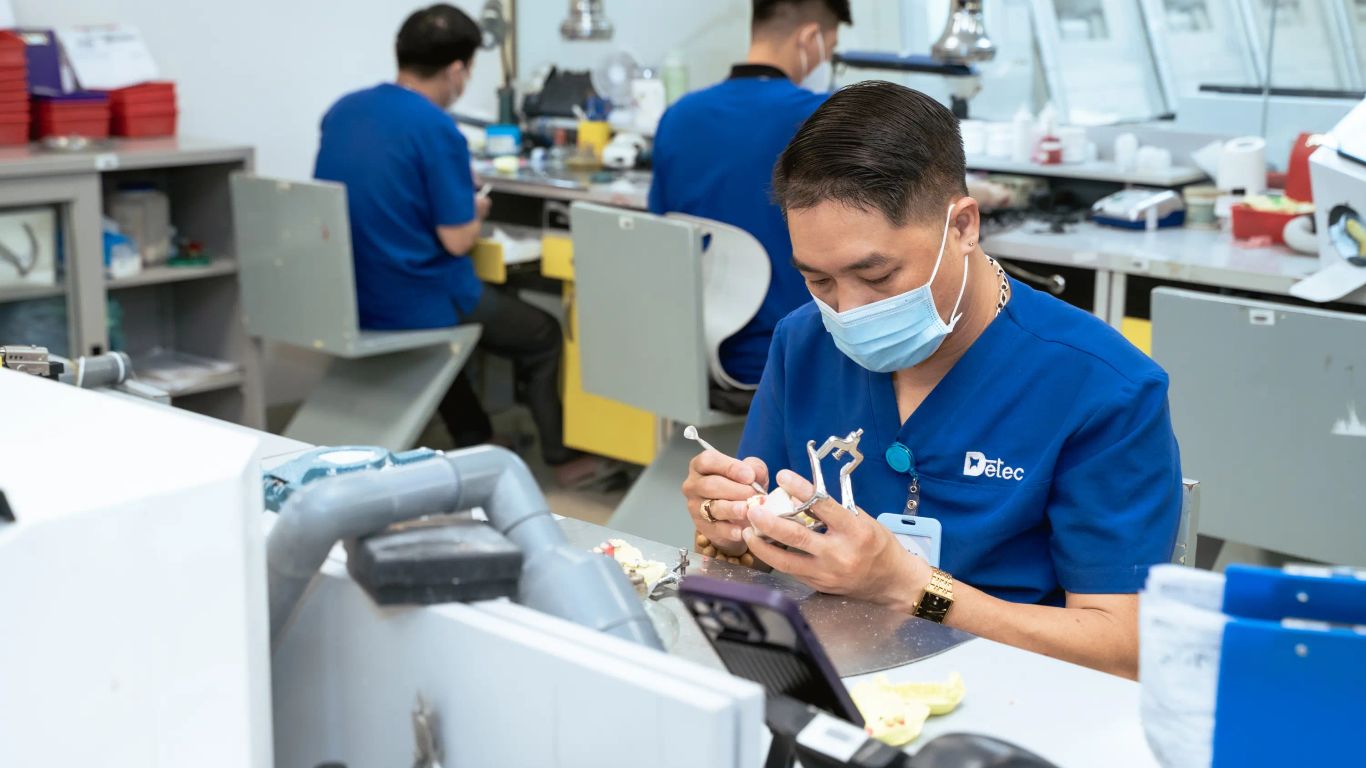 A person in a blue uniform works on a dental prosthesis in a lab.