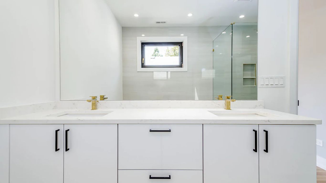 Modern white bathroom with double vanity, gold faucets, and glass shower.