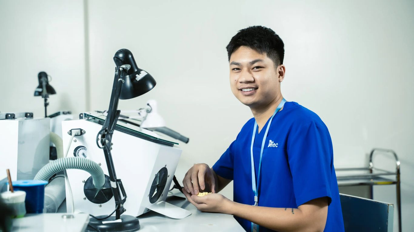A person in a blue uniform works with dental prosthetics at a lab bench.