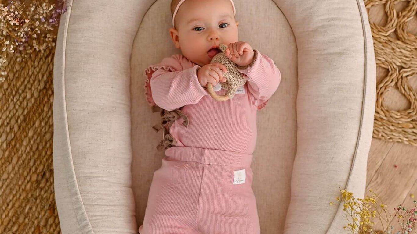 Baby in a pink outfit lies in a nest with a wooden toy.