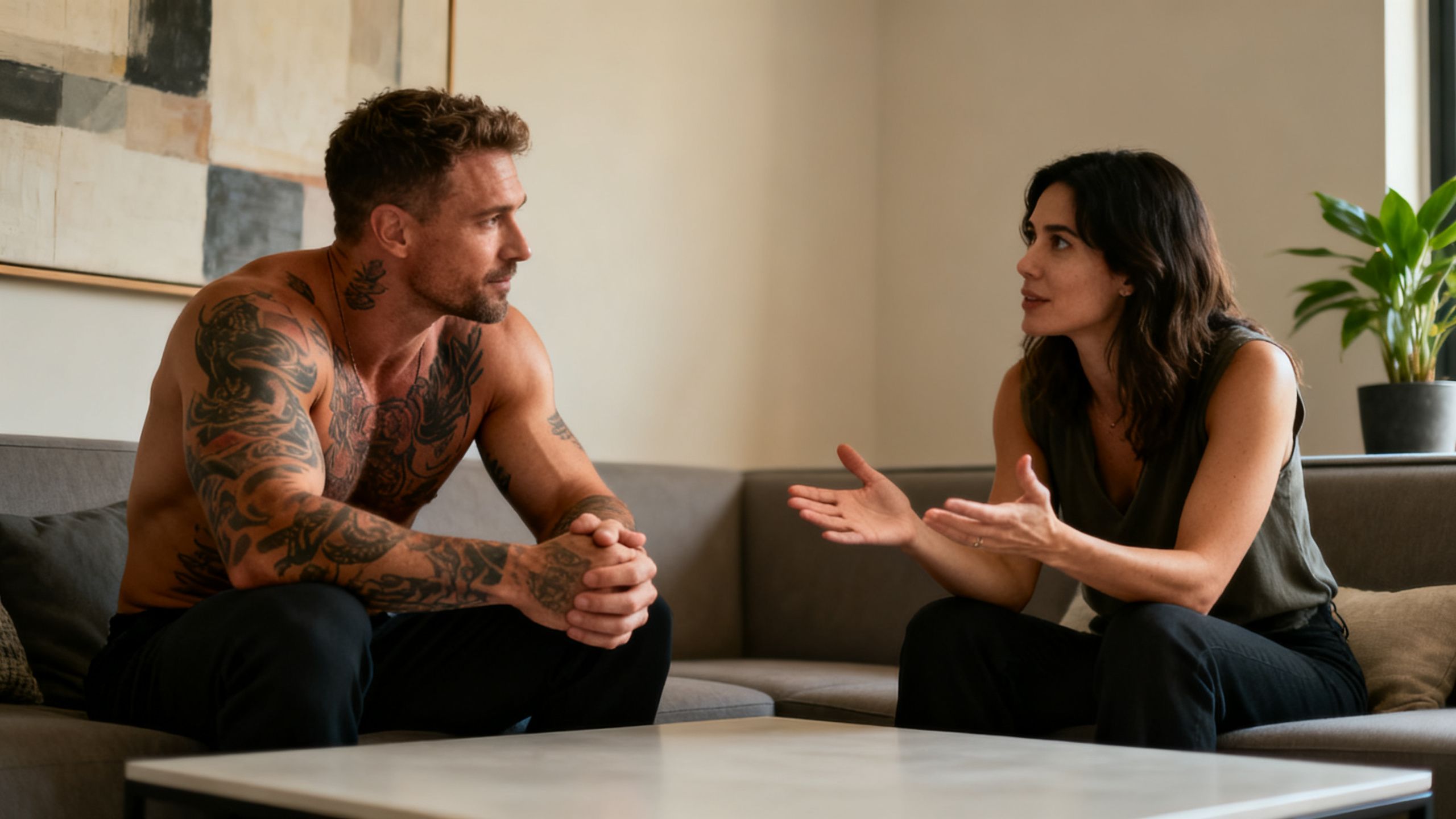 A muscular, tattooed man calmly talking with his partner in a modern living room, showing supportive communication.
