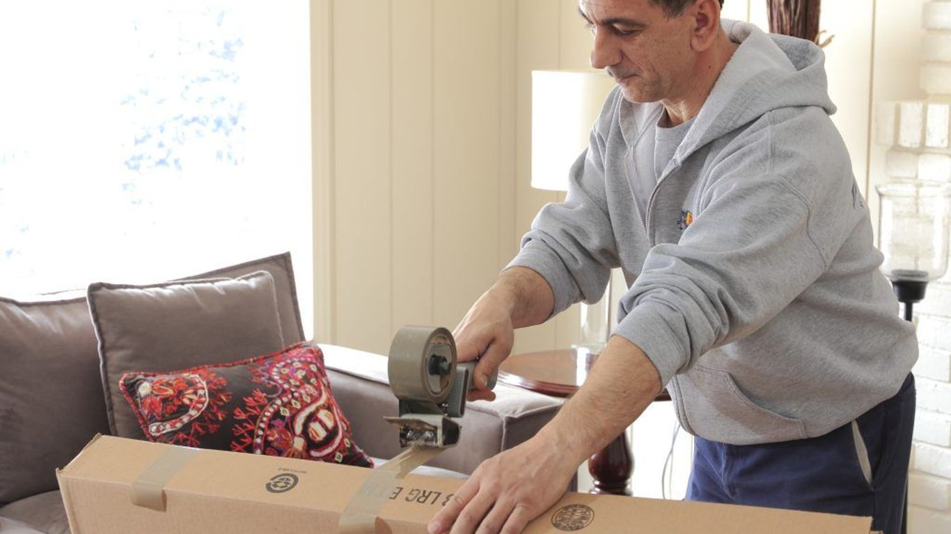 A person is taping a cardboard box shut in a living room.