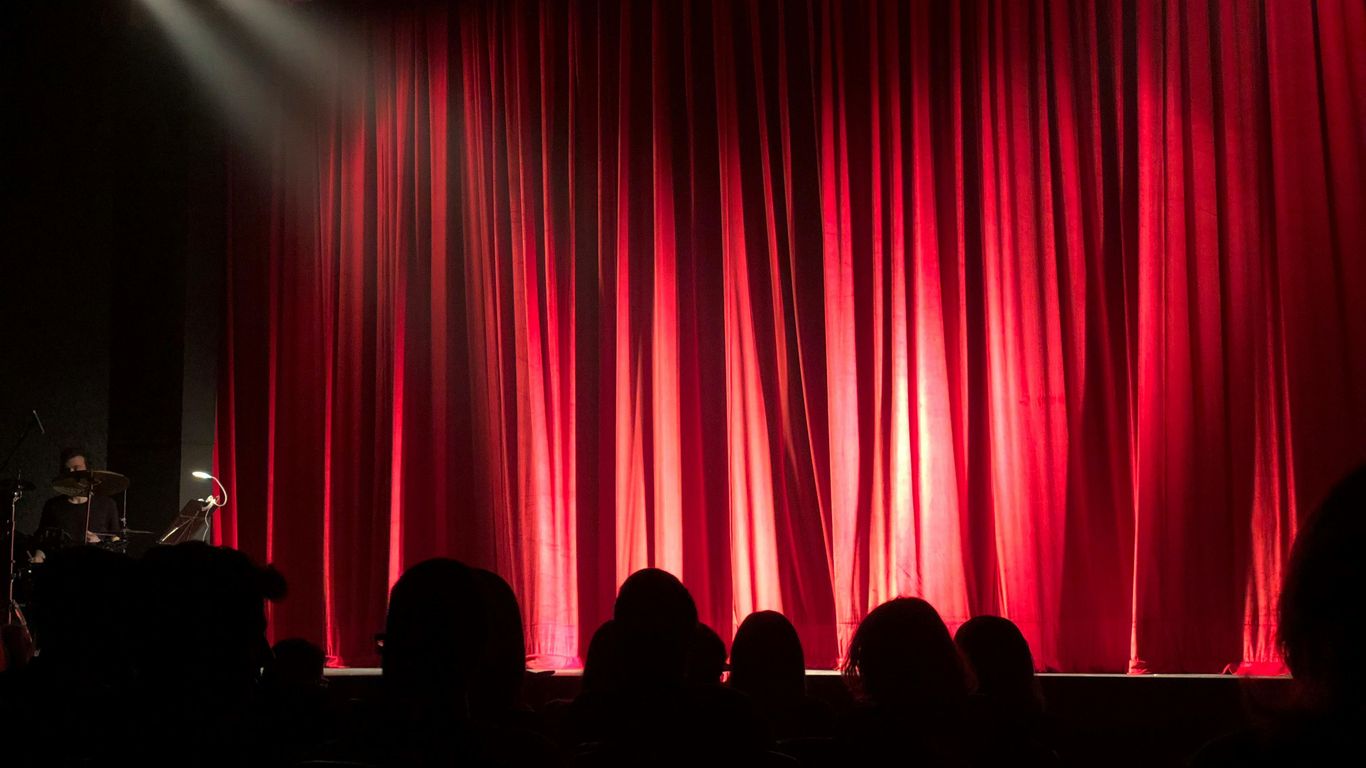 Theater stage with red curtains, audience in shadows waiting.