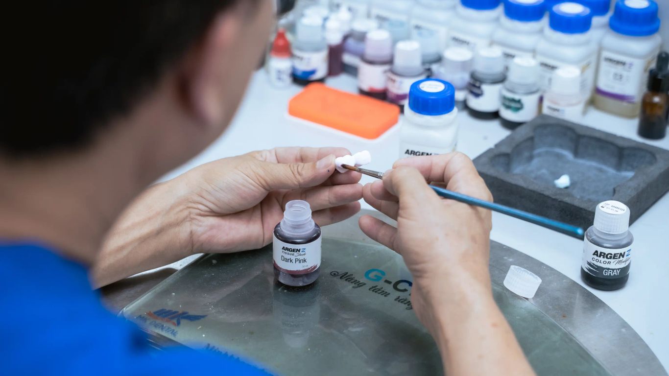 A person is painting a dental crown with pink and gray colors.