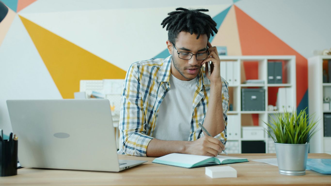 Hombre hablando por teléfono en el escritorio con laptop y libreta.