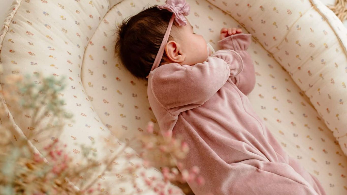 A baby sleeps peacefully in a floral-patterned nest with dried flowers.
