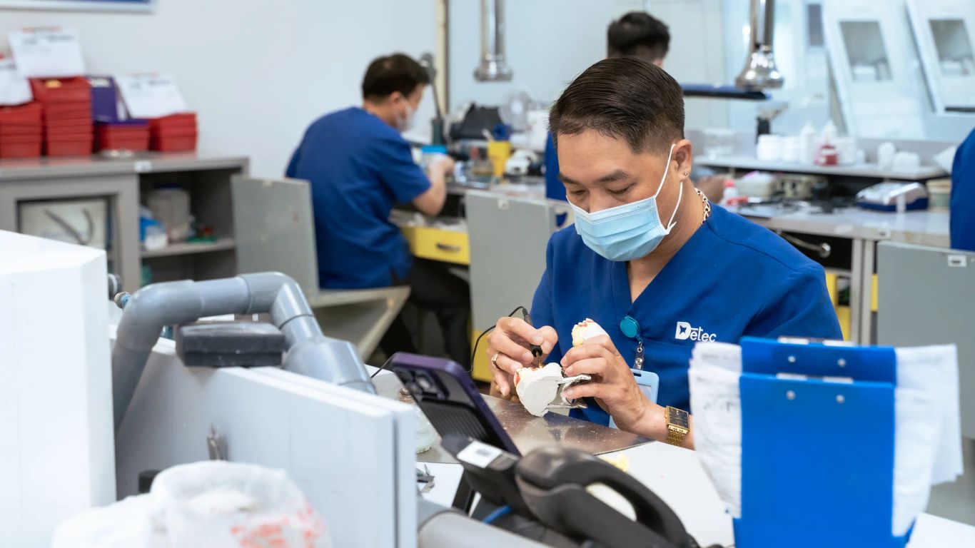 A person in a blue uniform works on a dental mold in a lab.