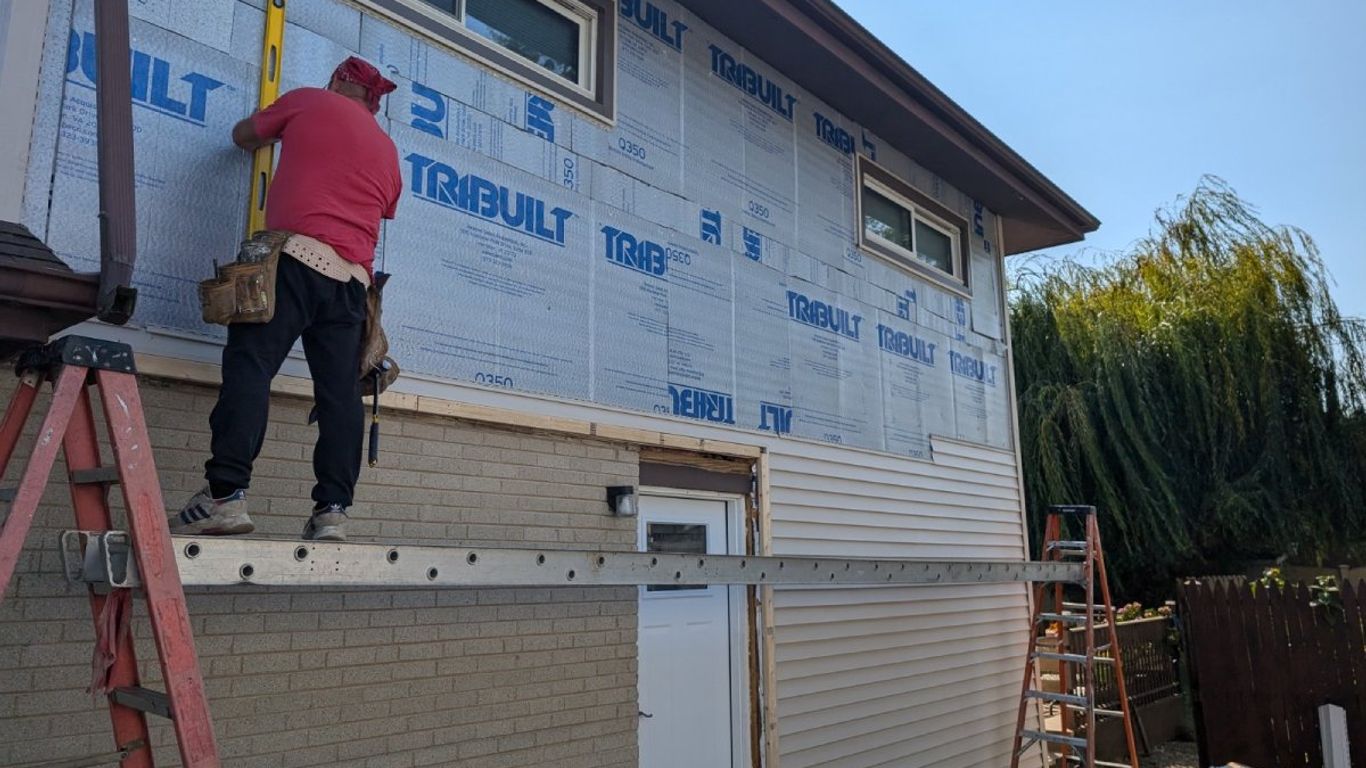 Construction worker installing siding on a house with ladders and tools.