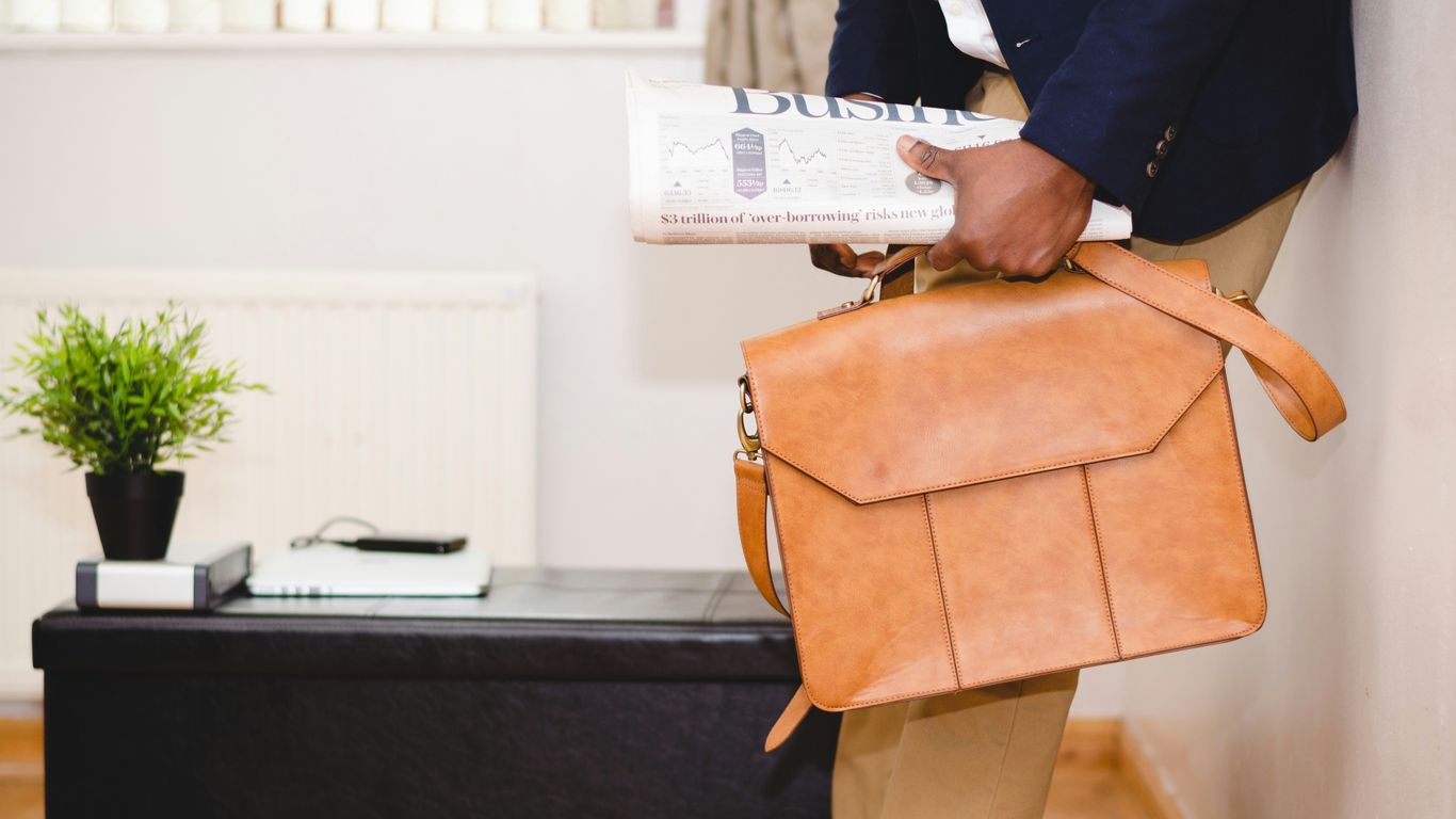 man holding brown leather bag leaning on white wall