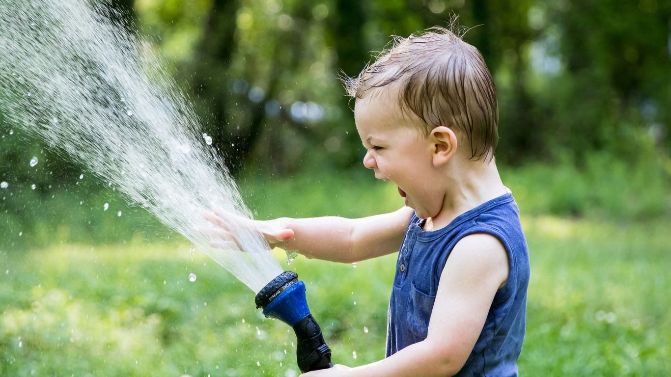 Child joyfully spraying water from a garden hose in a lush green yard, emphasizing outdoor fun and family-friendly activities.