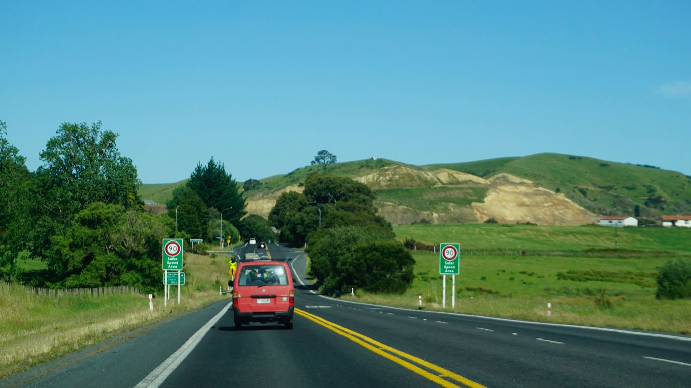 Red car driving on a highway through green hills.
