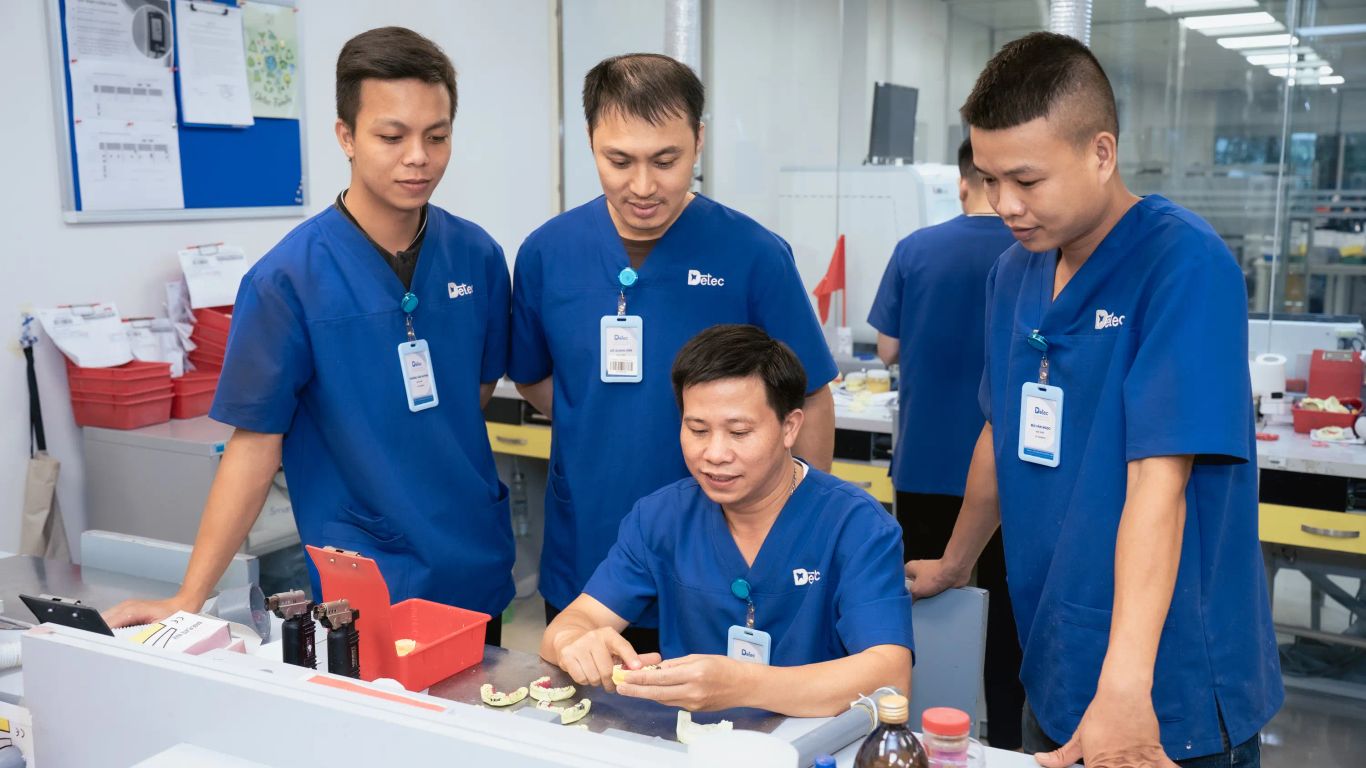 Workers in blue scrubs examine dental prosthetics in a laboratory.
