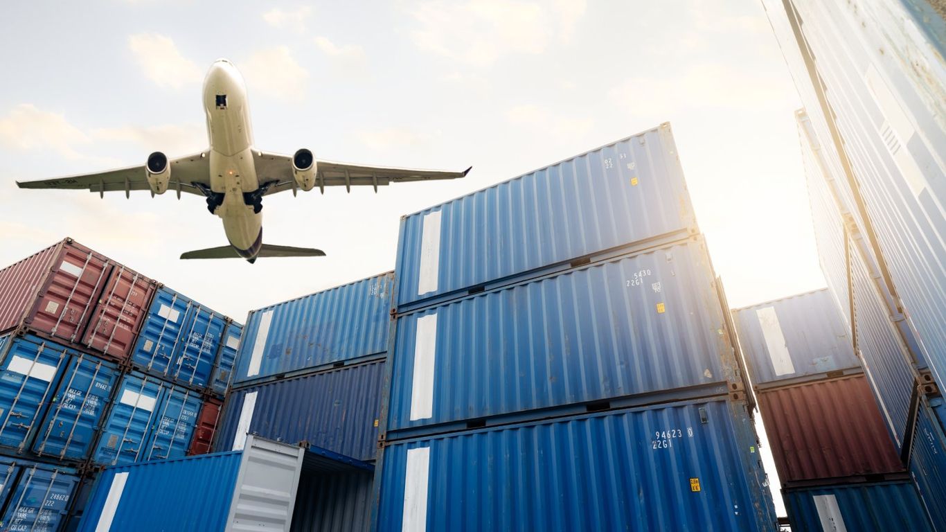 Airplane flying over stacks of shipping containers under a bright sky.