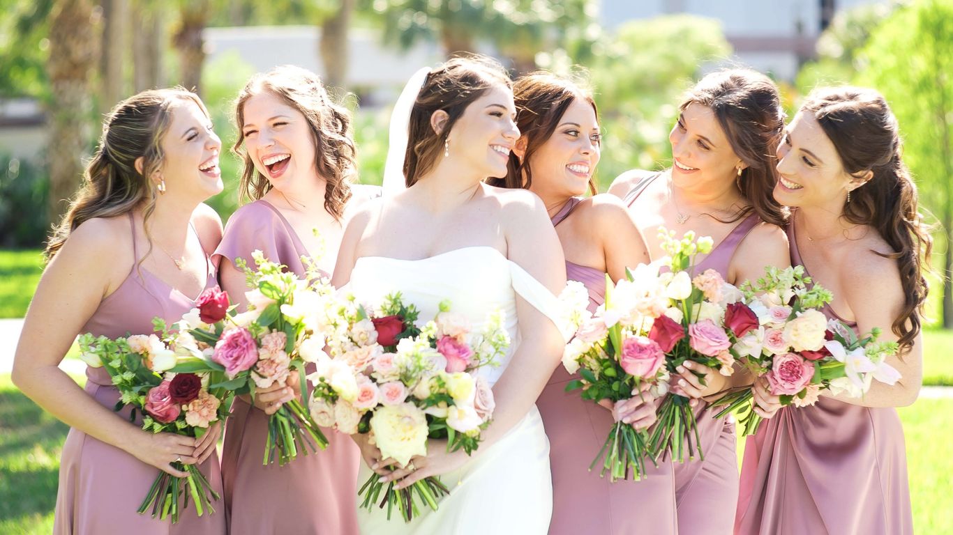 A bride and her bridesmaids laugh together holding bouquets.