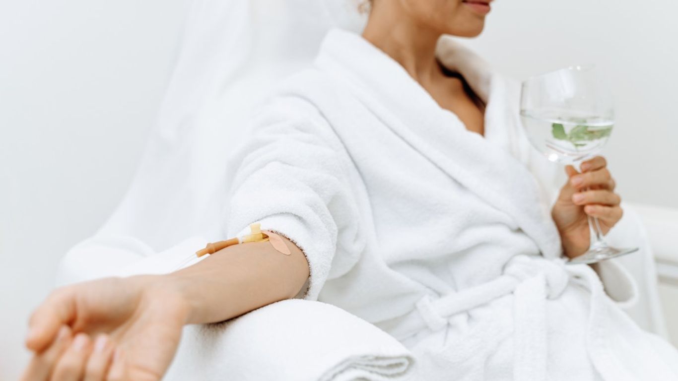 Person in a white robe receiving IV therapy while holding a drink.