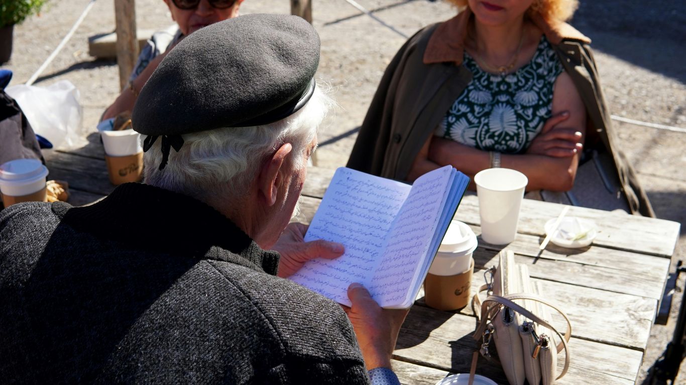 Man in beret reads notebook at outdoor table.