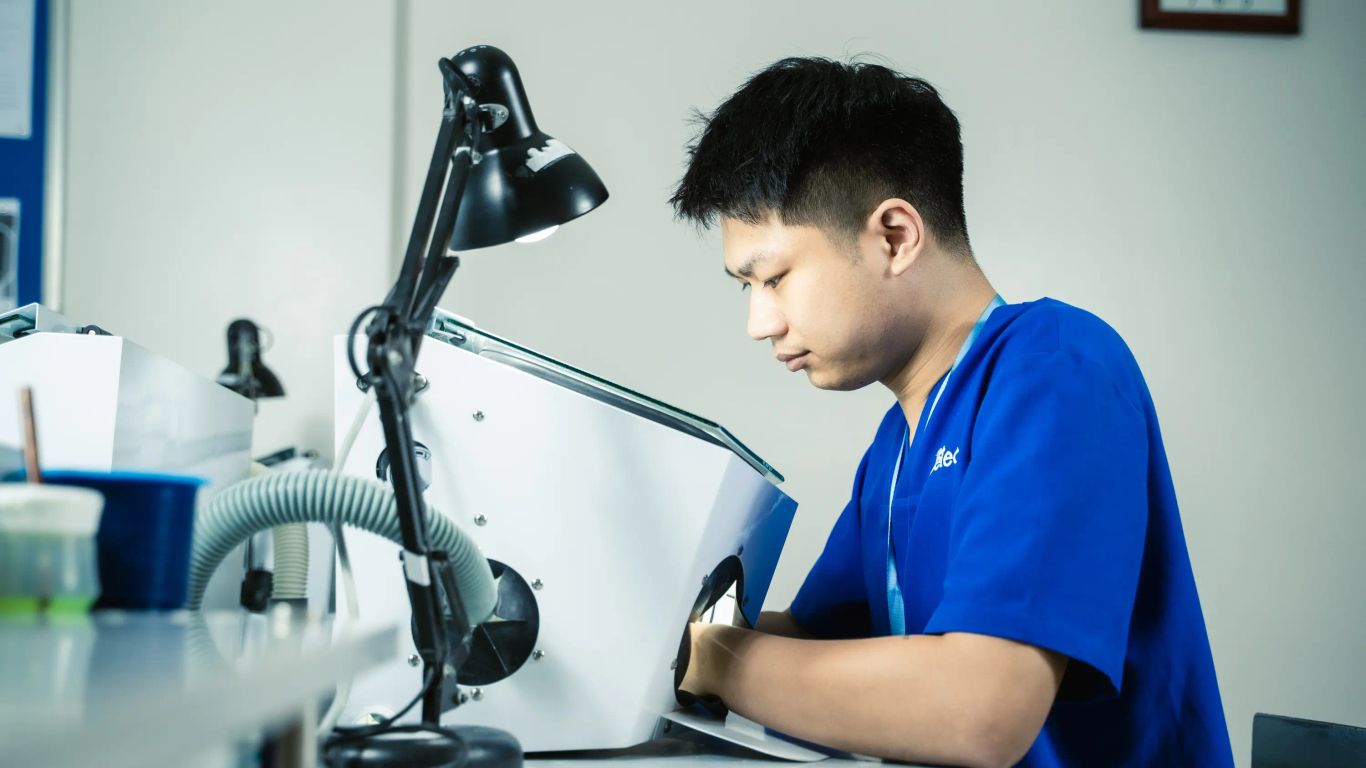 A person works intently with a dental lab machine under a bright lamp.