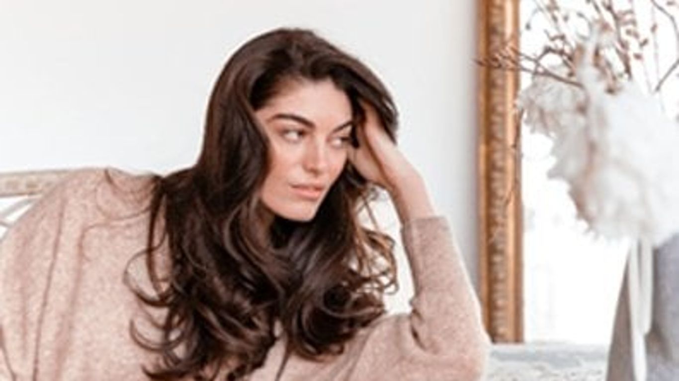 Woman with long hair sitting indoors, looking thoughtful or distracted.