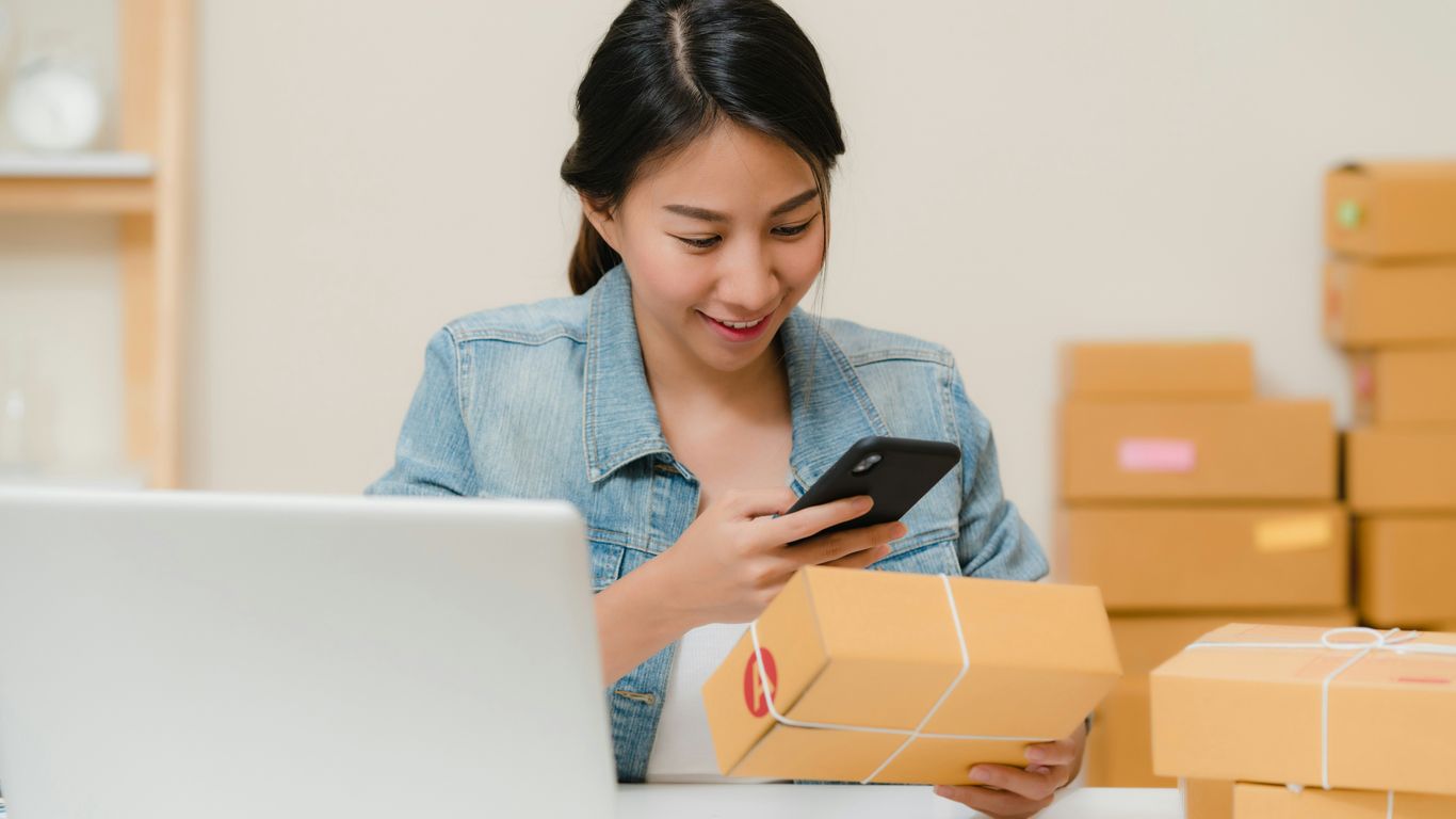 Woman checking package with phone near laptop and boxes