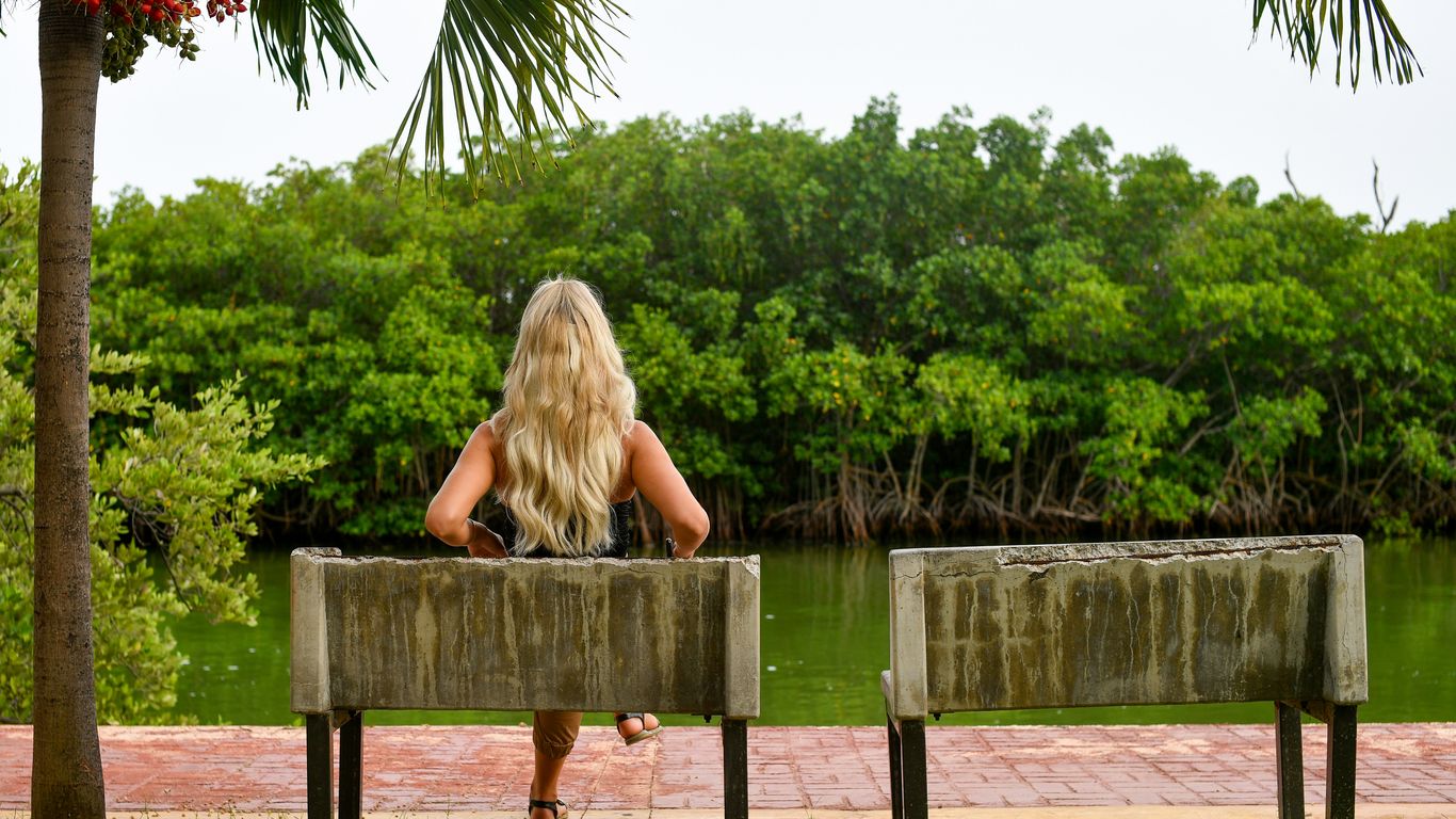 Woman with blonde hair sits on a bench overlooking water.