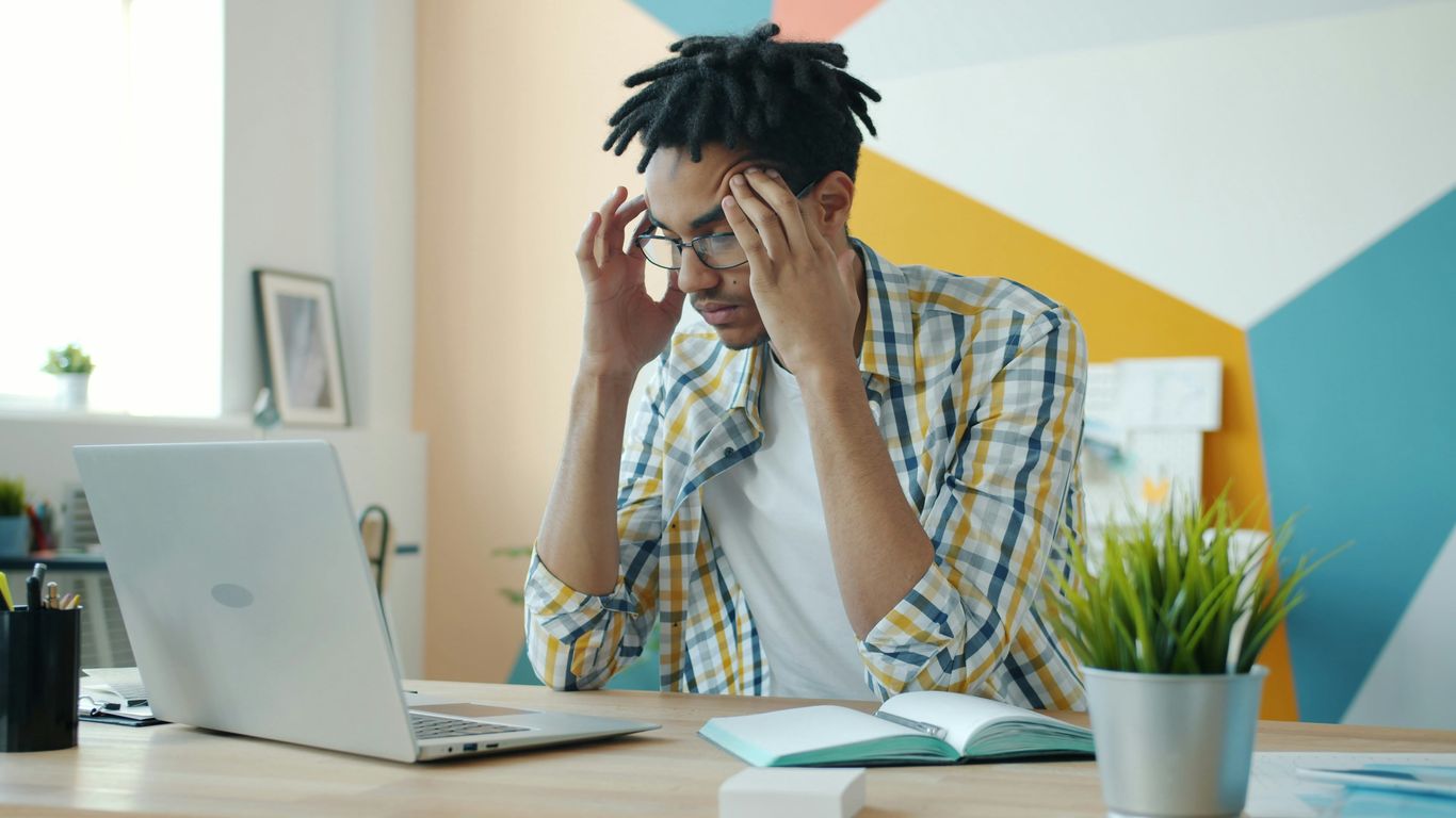 Man with dreadlocks holding head at desk with laptop