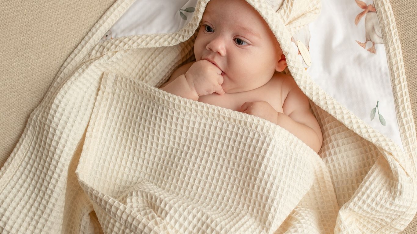 A baby wrapped in a cream-colored waffle knit blanket with a duck print hood.