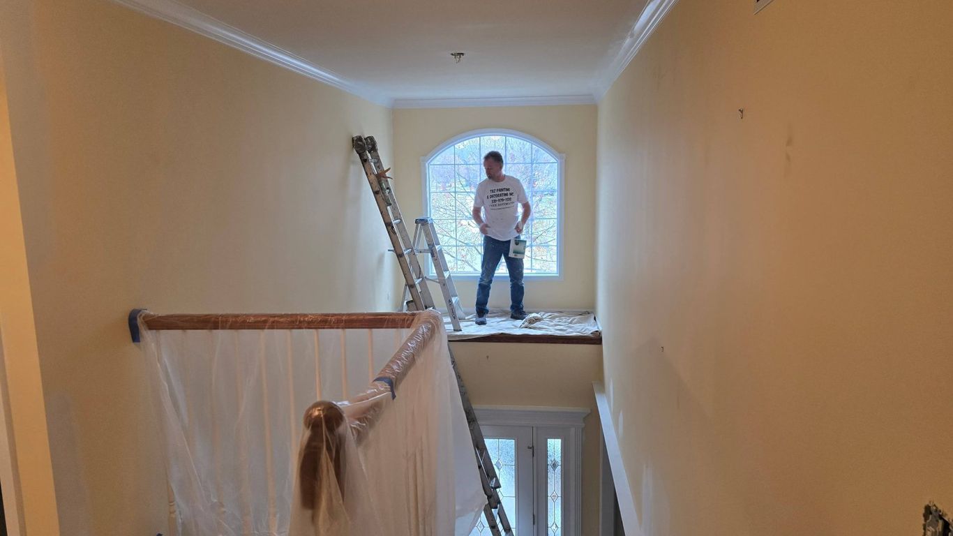 A person stands on a ladder with a paint can near a window.