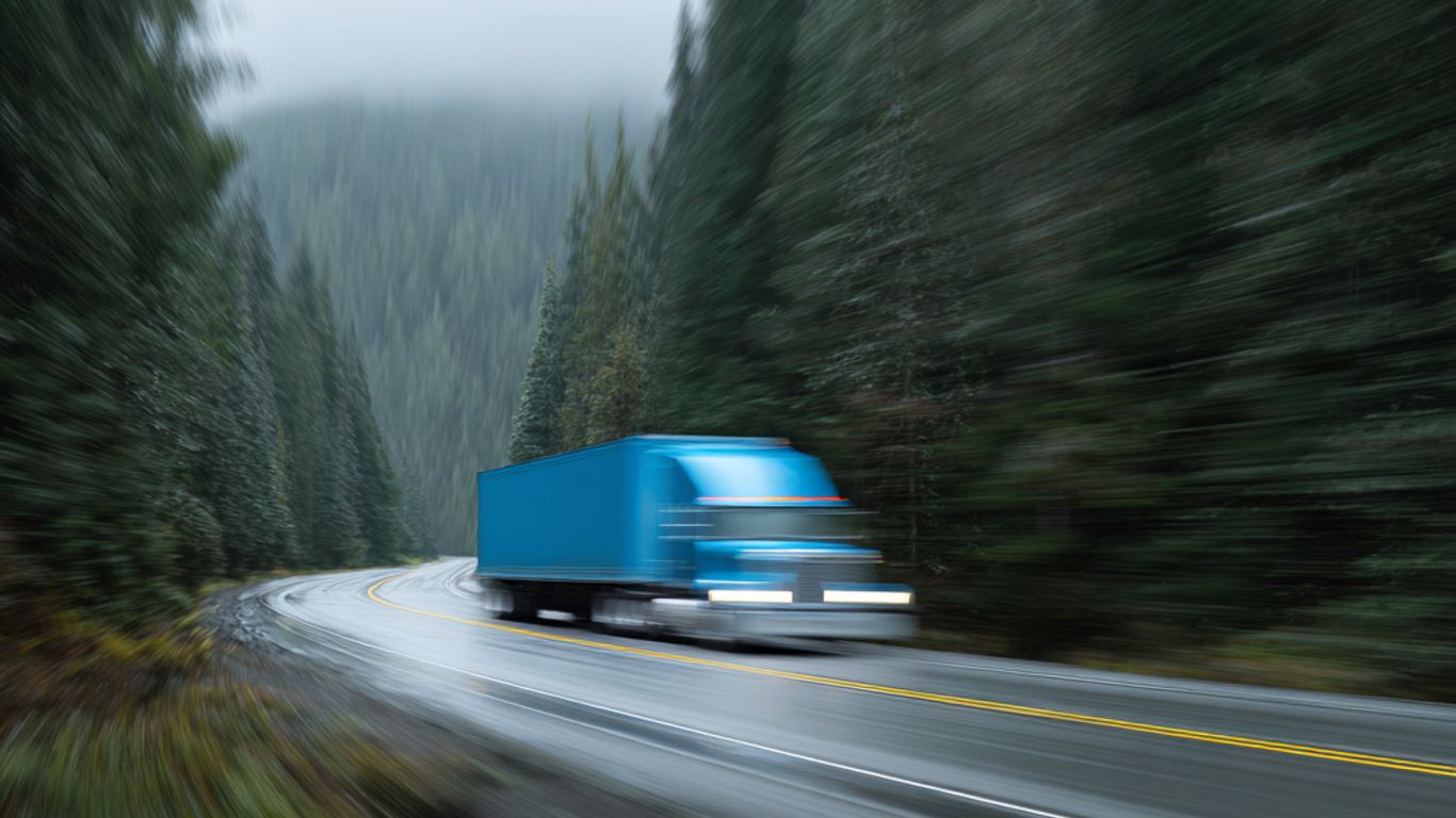 A blue semi-truck speeds down a wet, winding road through a forest.
