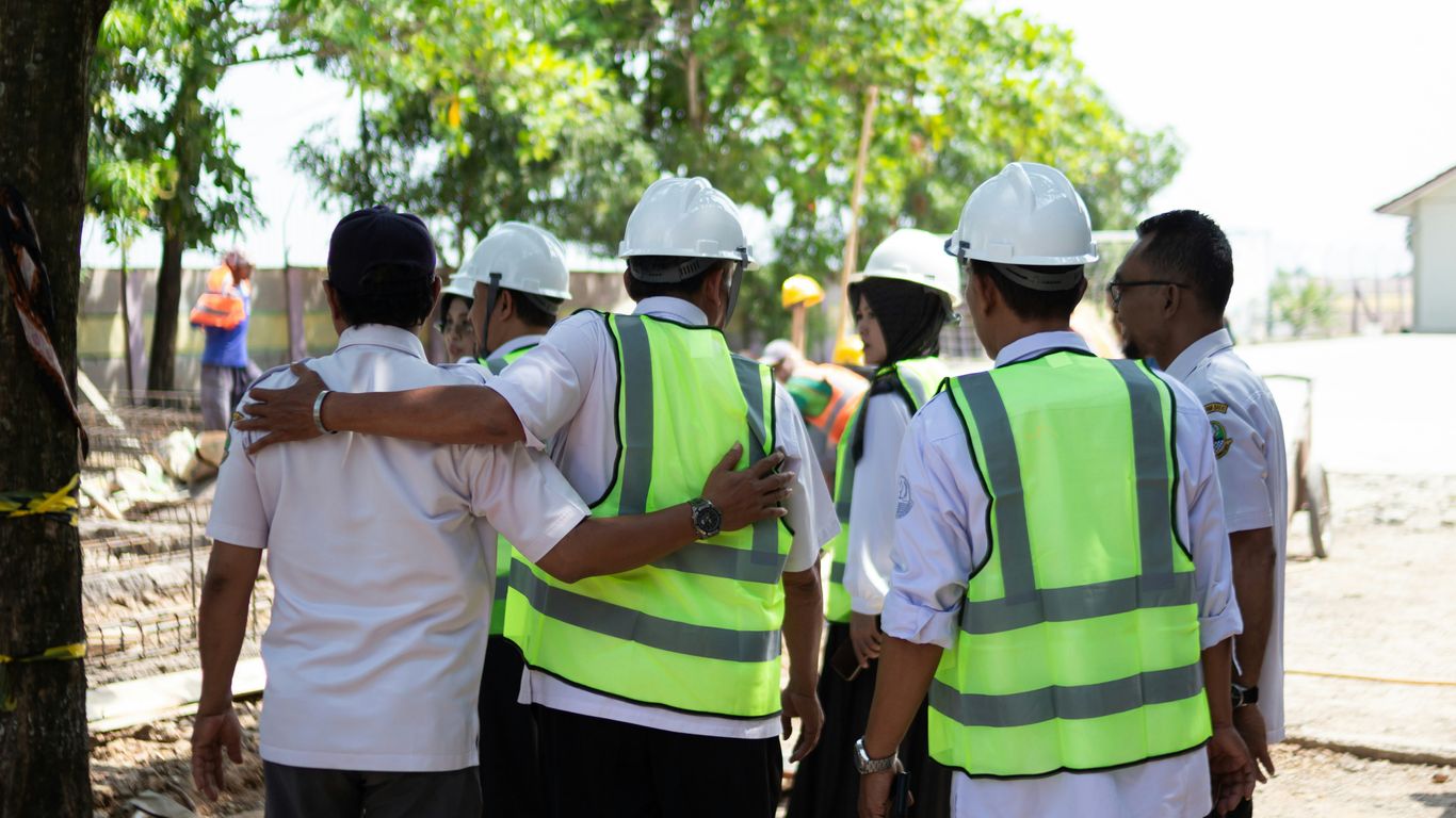 Construction workers in hard hats and vests huddle together.