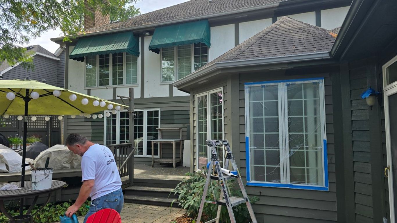 A person is watering plants near a house being painted.