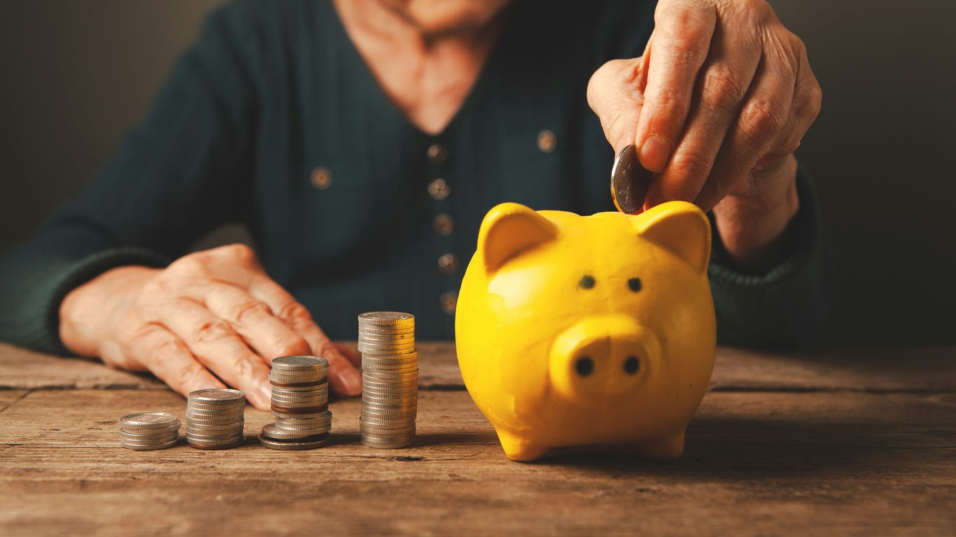 Elderly hands depositing coins into a yellow piggy bank.