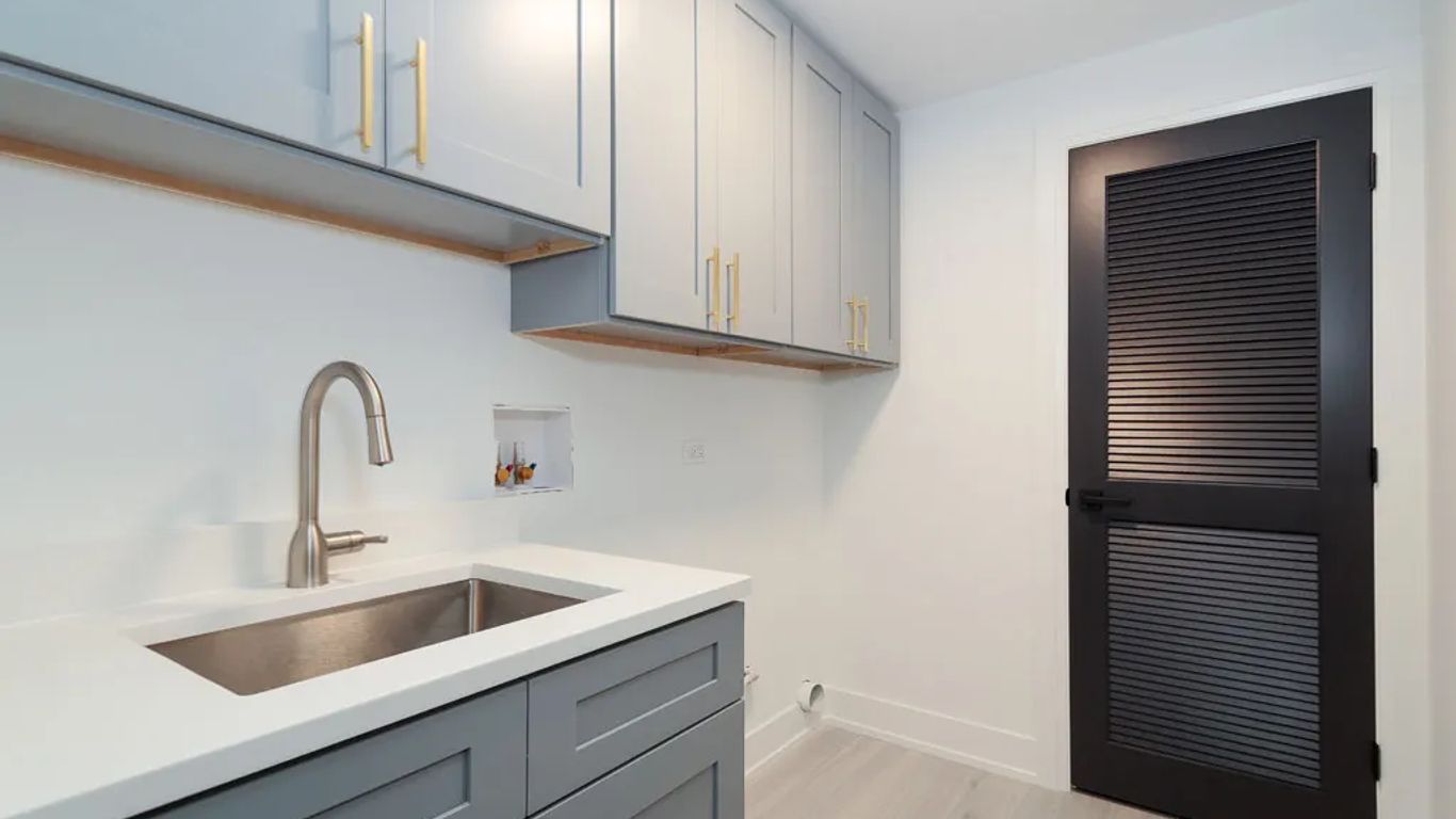 A modern laundry room with gray cabinets, a stainless steel sink, and a black door.