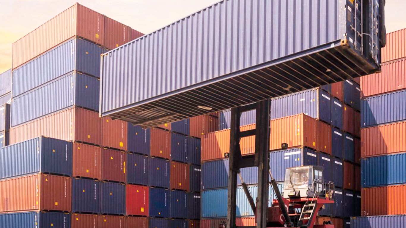 A forklift lifts a shipping container among stacks of containers.