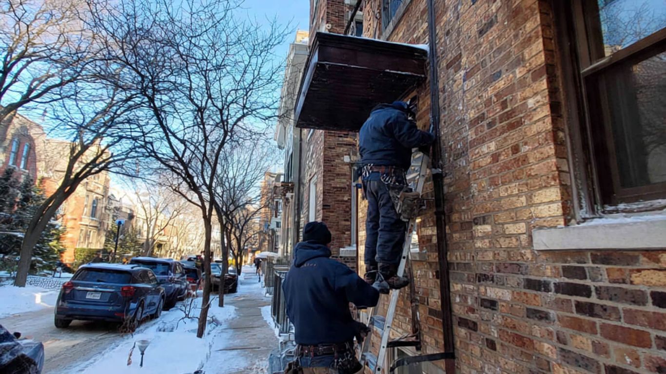 Workers on ladders perform repairs on a brick building in the snow.