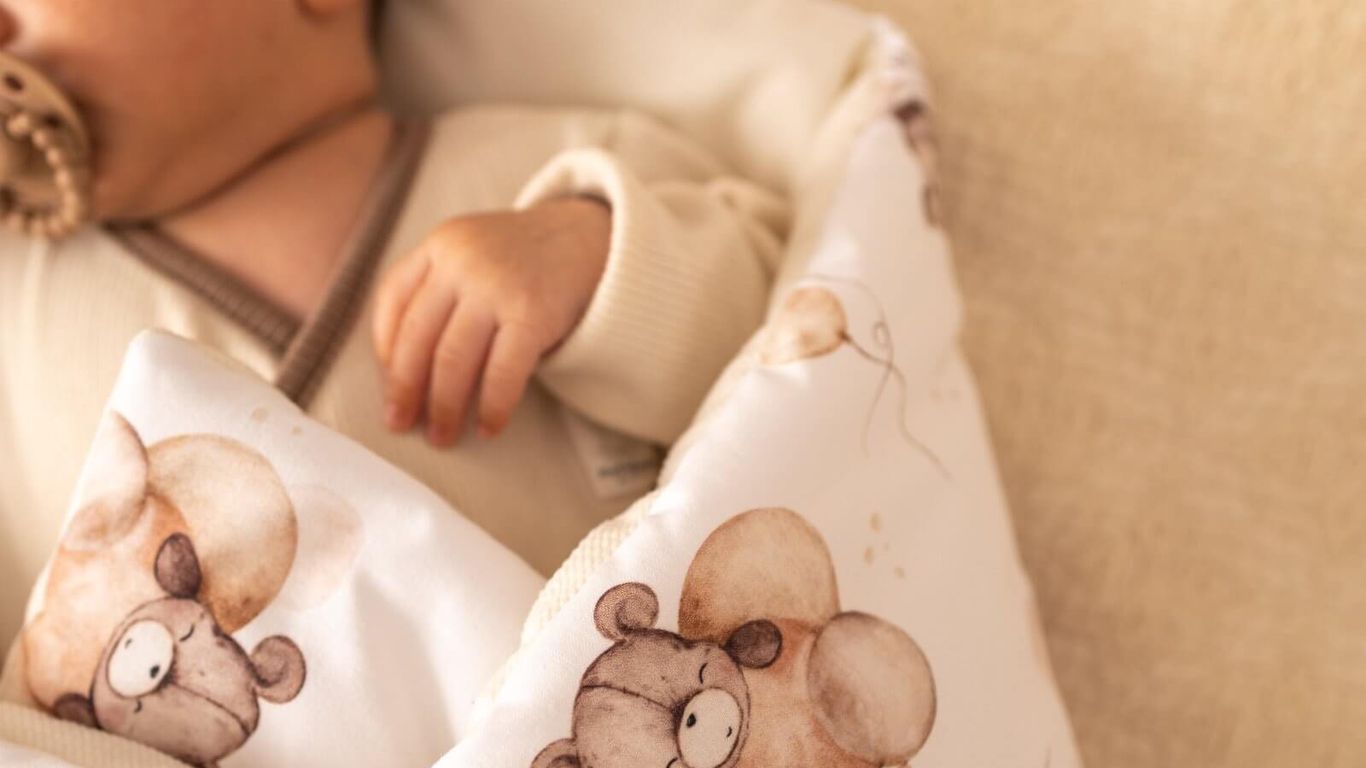 A baby sleeps peacefully wrapped in a blanket with teddy bear and balloon print.