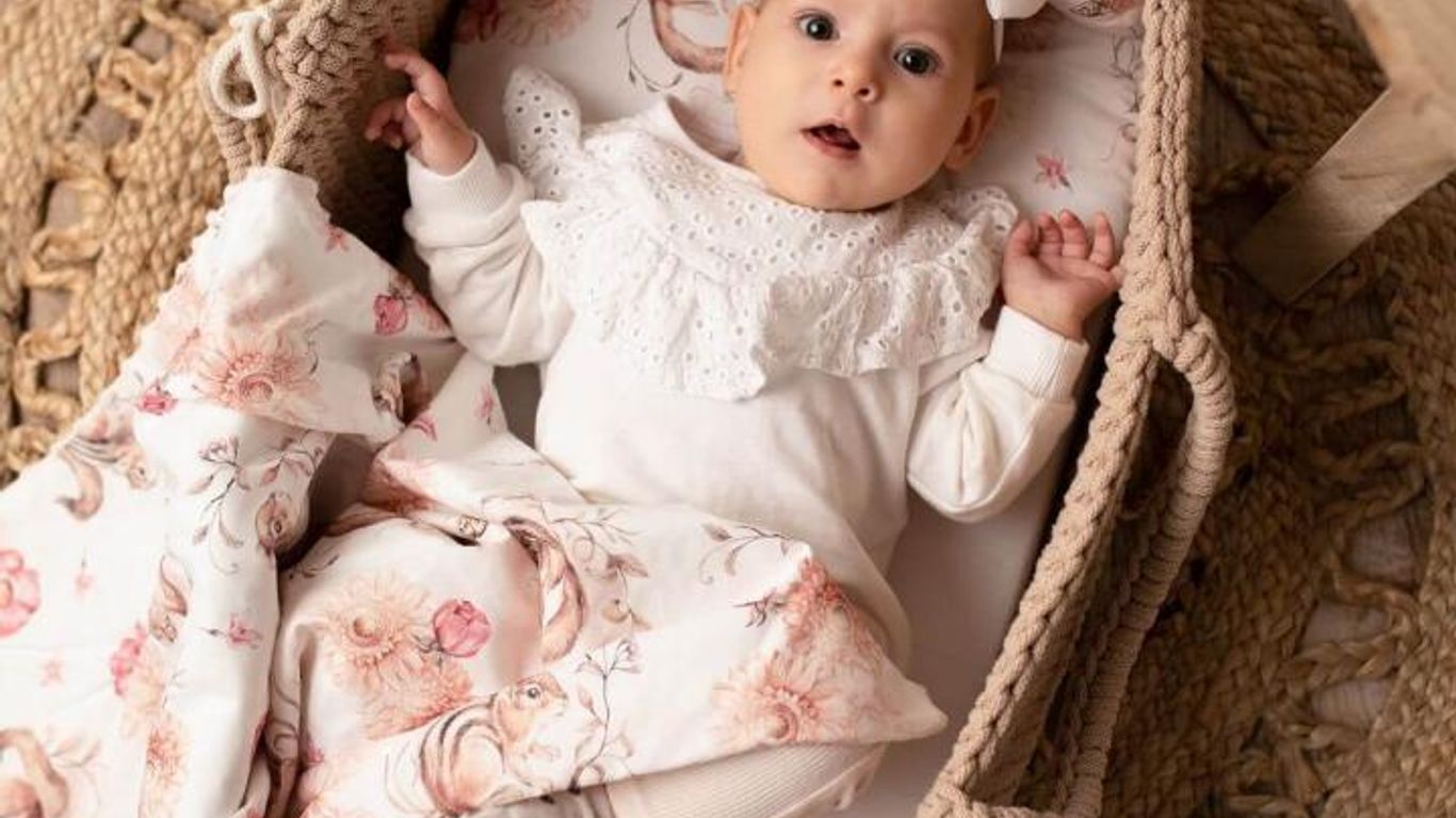 A baby lies in a bassinet with a floral blanket and headband.