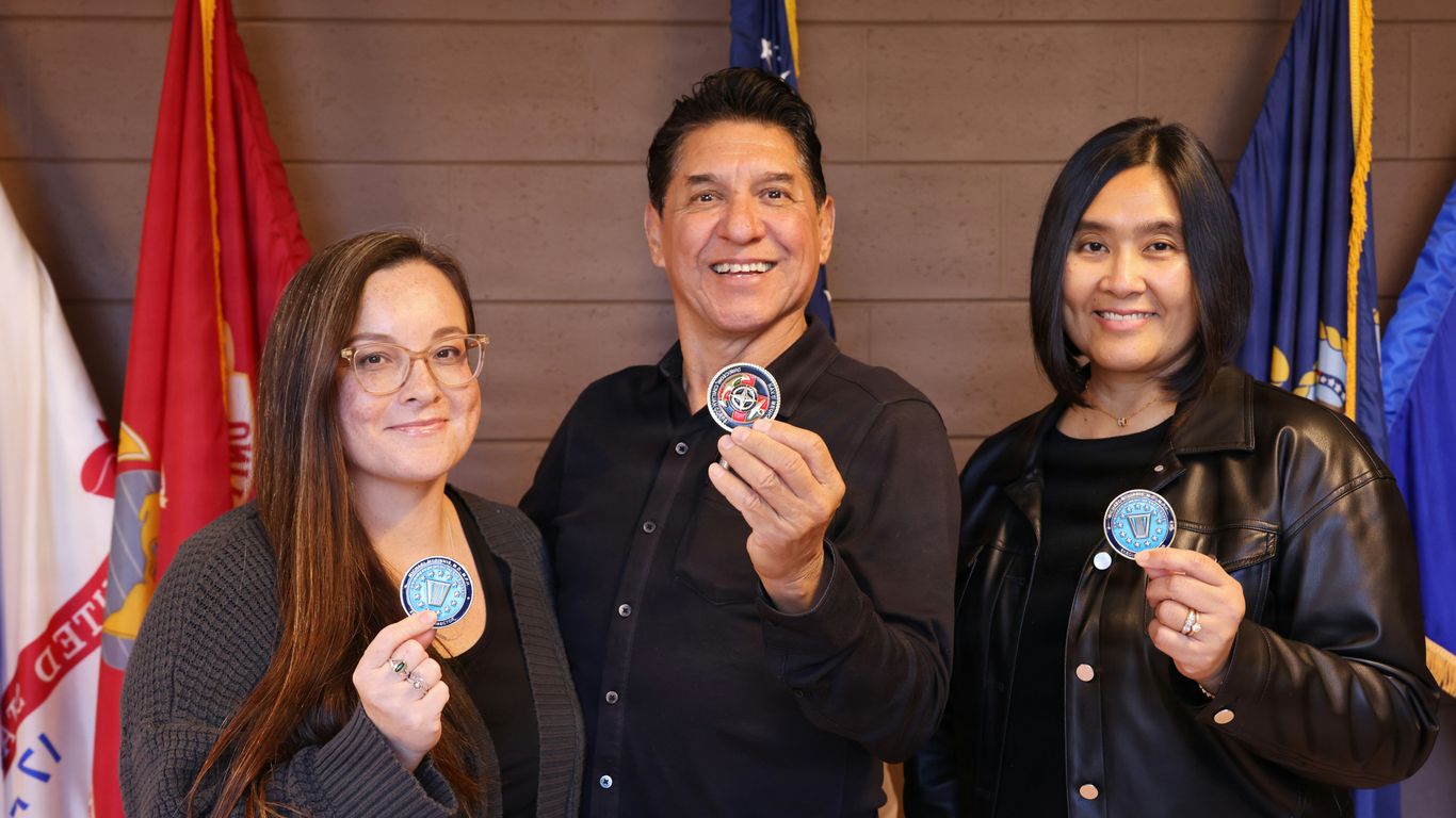 Three people hold up medals in front of flags