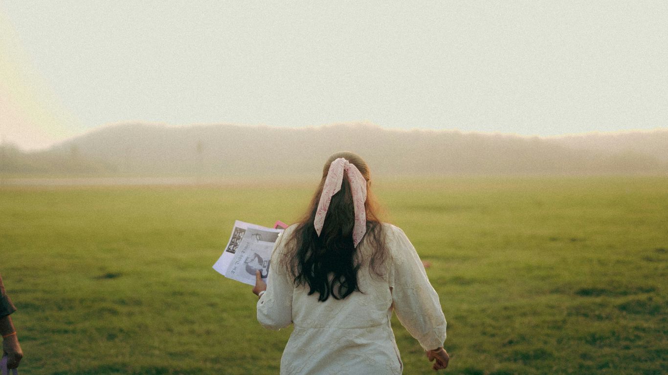 Woman holding newspaper walks through a grassy field.