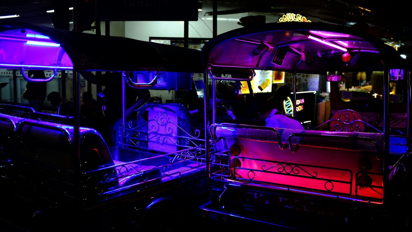 Two tuk-tuks illuminated by colorful neon lights at night.