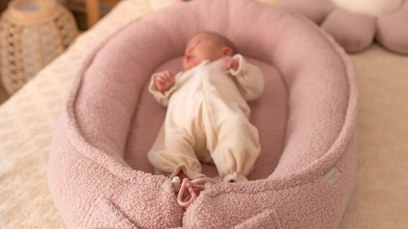 A baby sleeps in a pink fuzzy nest with a bow.