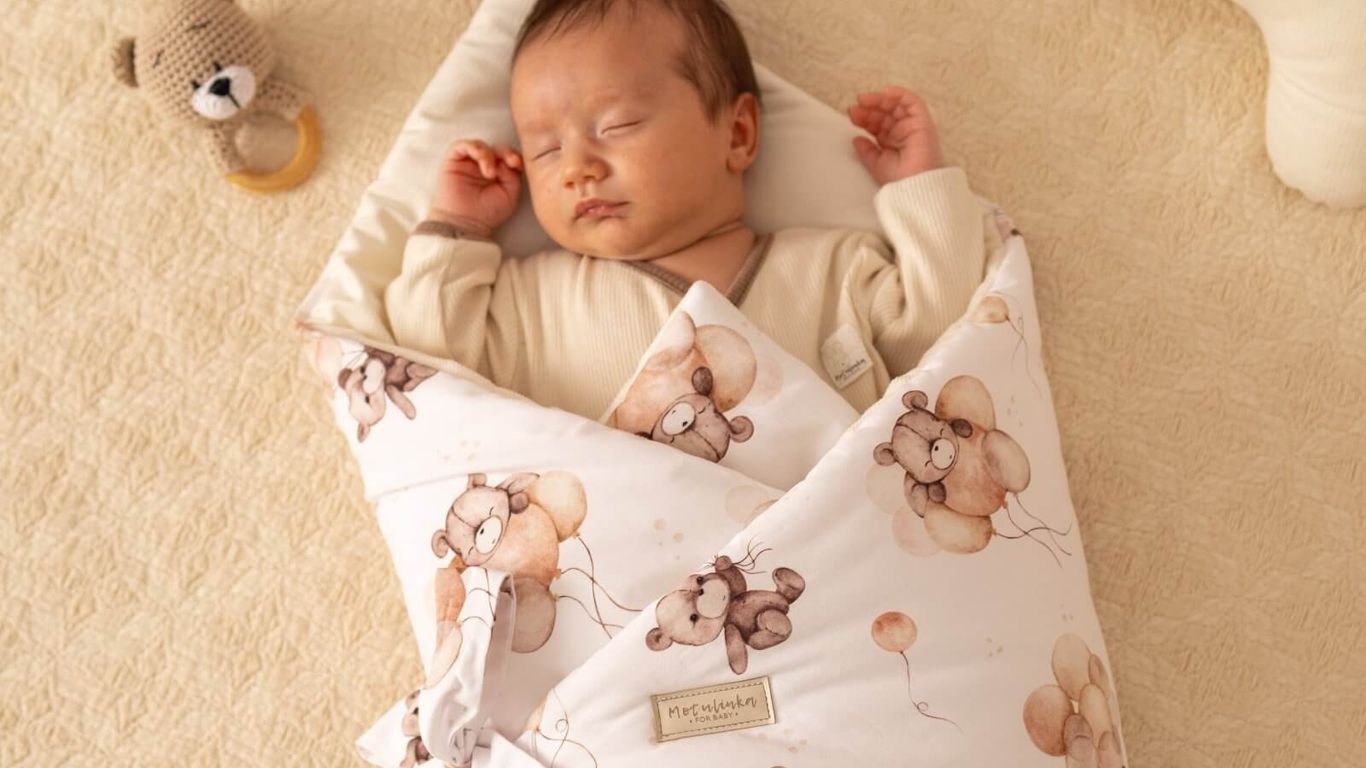 A baby sleeps peacefully wrapped in a teddy bear patterned swaddle.