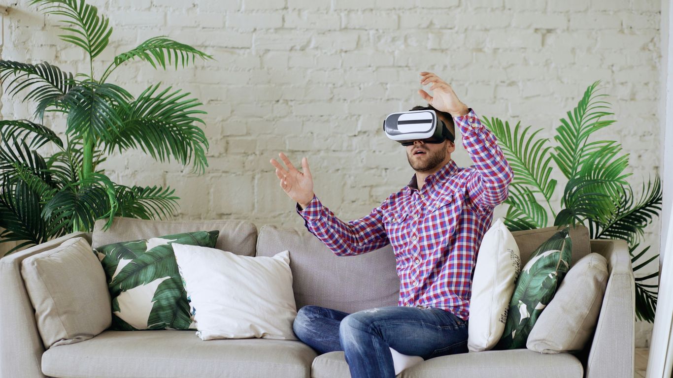 Man wearing vr headset sitting on couch, gesturing with hands.
