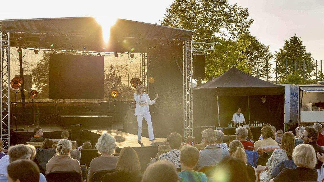A performer sings on stage to an outdoor audience at sunset.