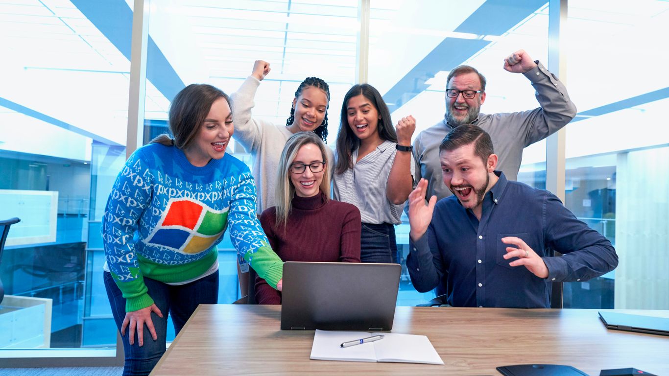 men and women sitting and standing by the table looking happy while staring at laptop