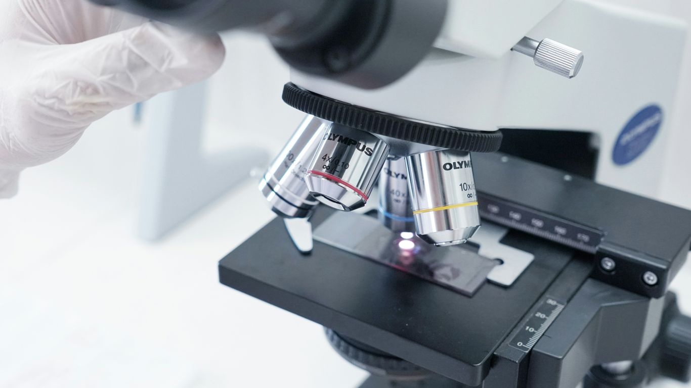 A person in white gloves is looking through a microscope