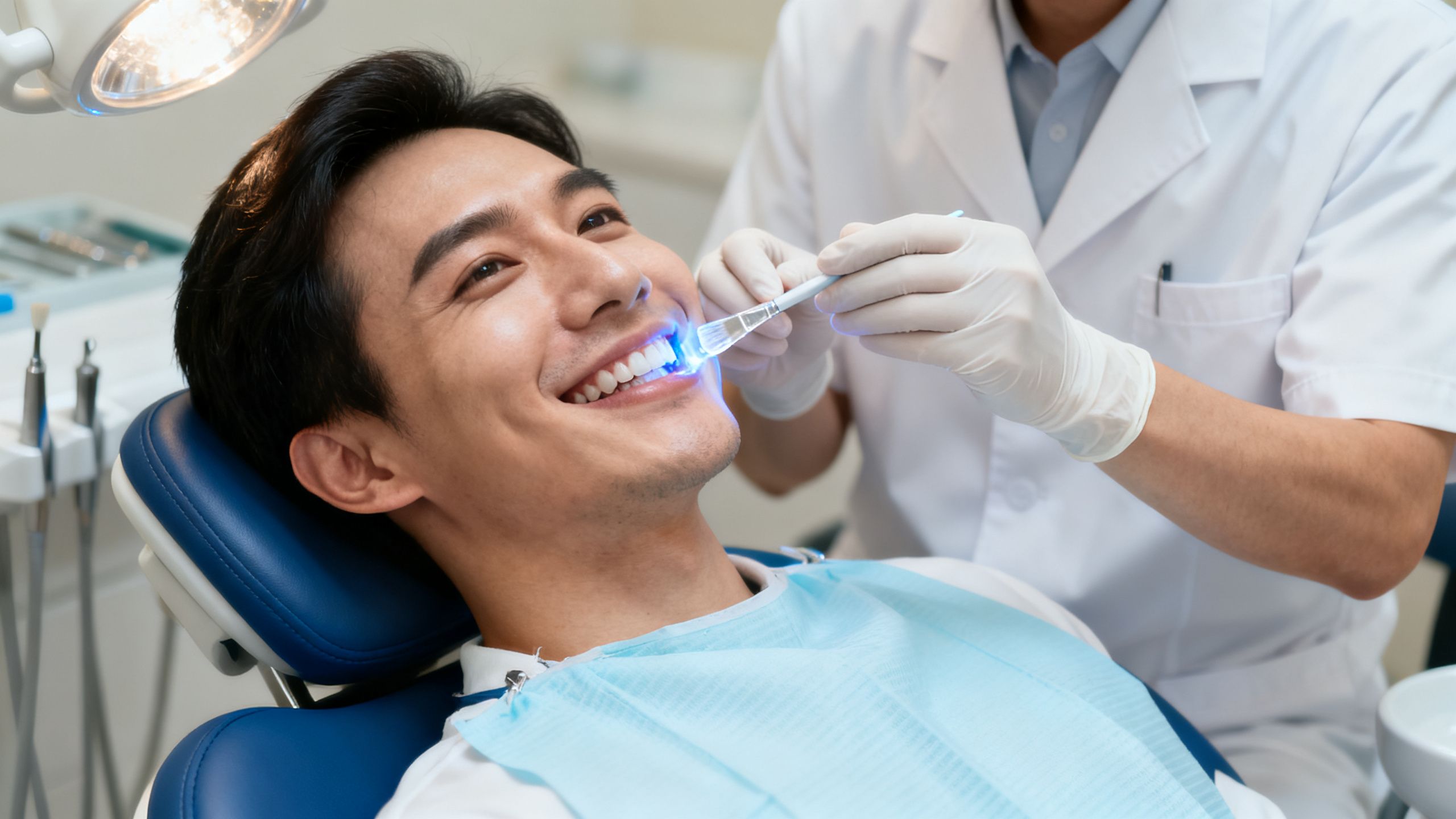 A patient smiling in a dental chair while a dentist applies a whitening gel to their teeth.