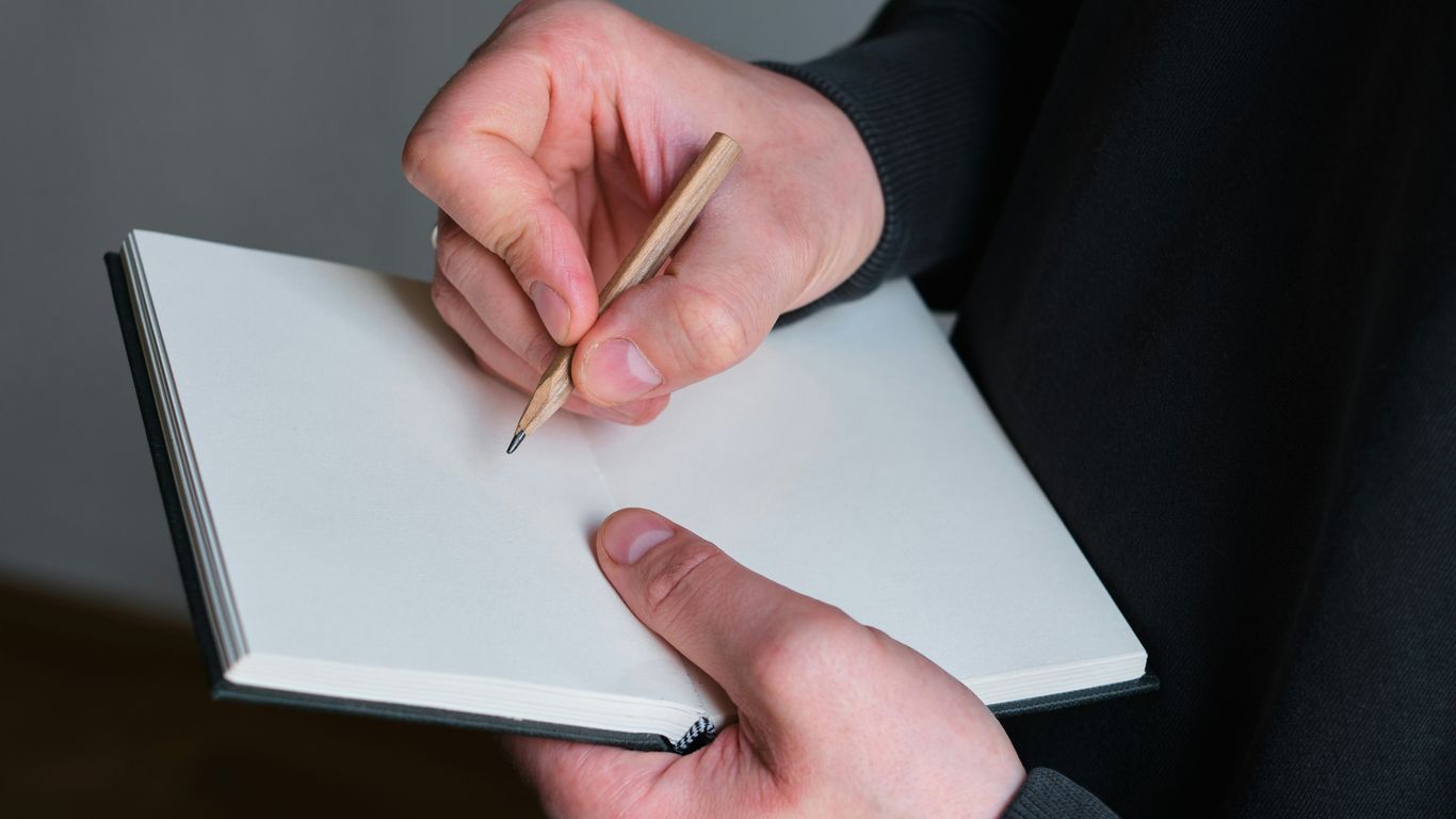 Person writing in a blank notebook with pencil.