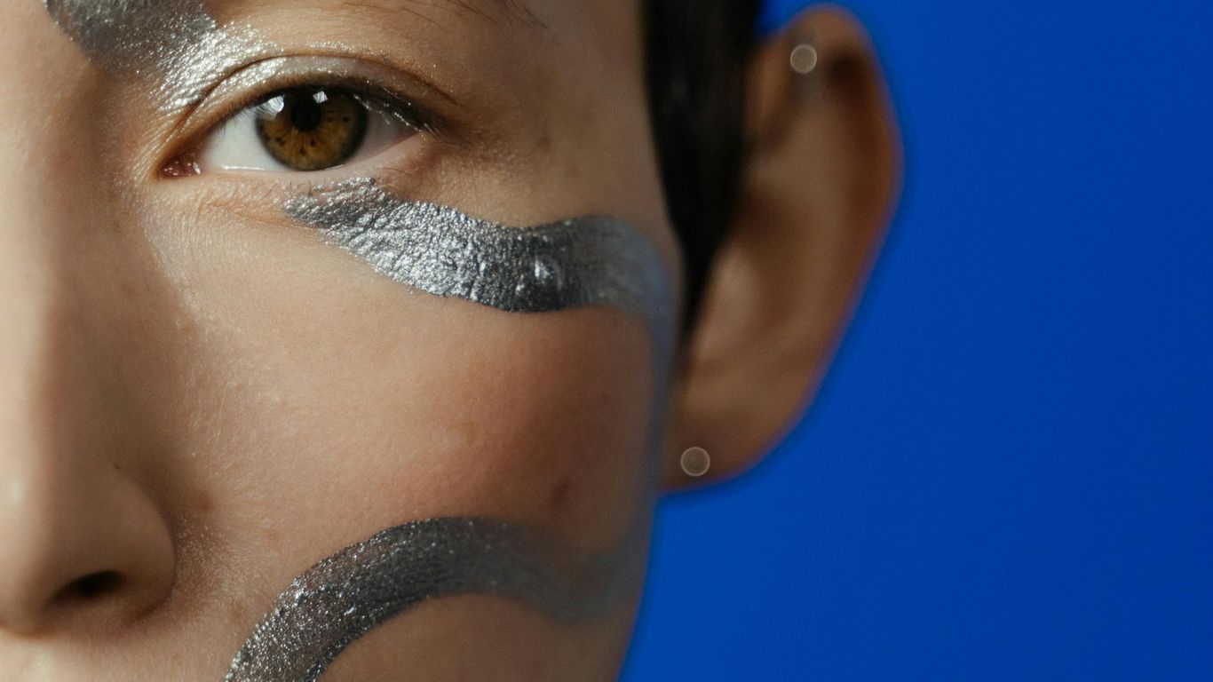 Close-up of a face with silver body paint against a blue background.
