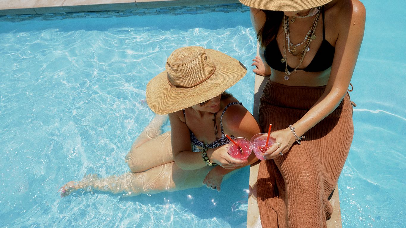Two people in wide-brimmed hats enjoy drinks by a swimming pool.