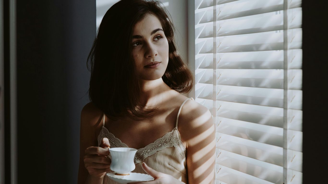 Person holding teacup, standing by window with blinds, thoughtful.